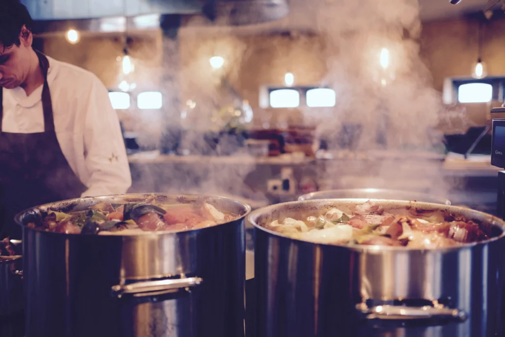 Smoke piles up in the kitchen as a man is at work wearing a black apron near two silver metal smoking cooking pots.