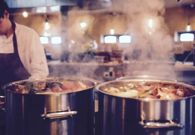 Smoke piles up in the kitchen as a man is at work wearing a black apron near two silver metal smoking cooking pots.